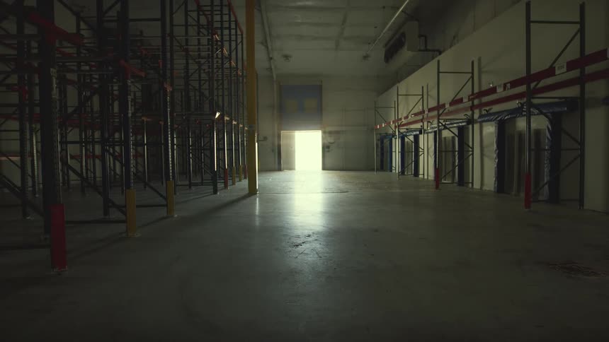 A wide, static view inside a large, desolate, and empty warehouse with shelving units and a bright light at the end