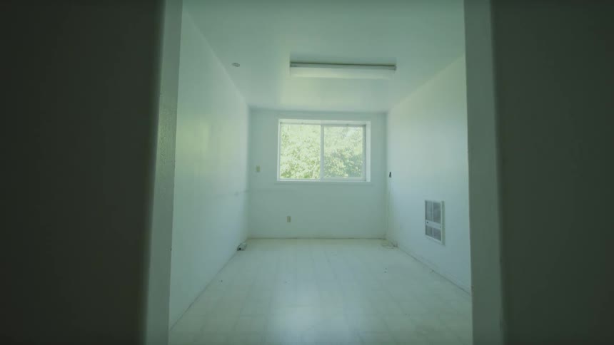 Slowly backing away from a dark wall to reveal an empty, unfurnished room with white walls and a window