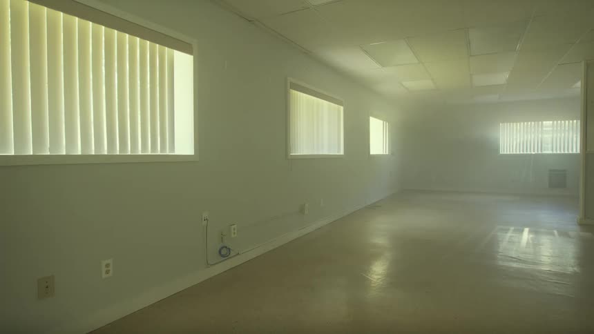A long, empty room is filled with hazy sunlight pouring through the vertical blinds on the windows