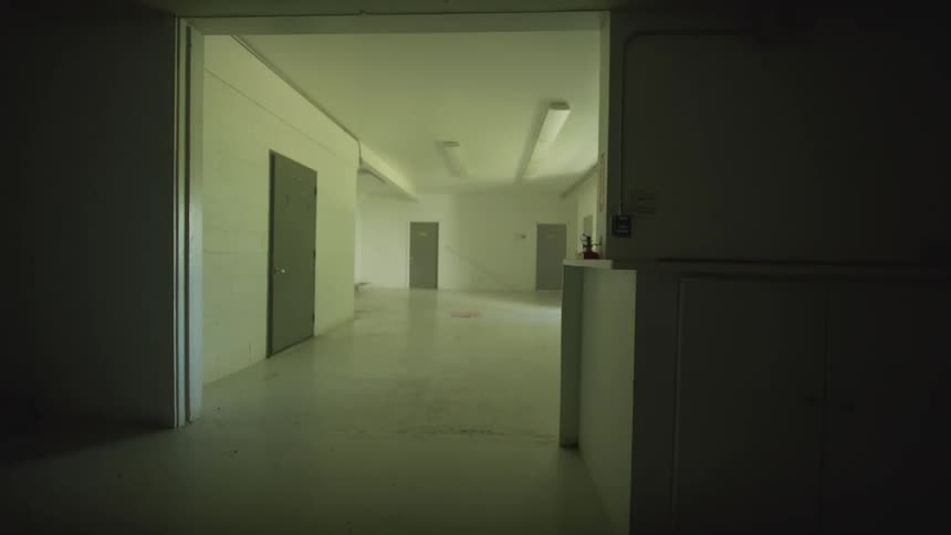 An unsettling view from a dark room into a brightly lit but empty institutional hallway with flickering fluorescent lights