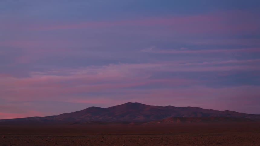 A stunning timelapse of a vibrant pink and purple sunset over a desert mountain range as clouds drift by and night falls