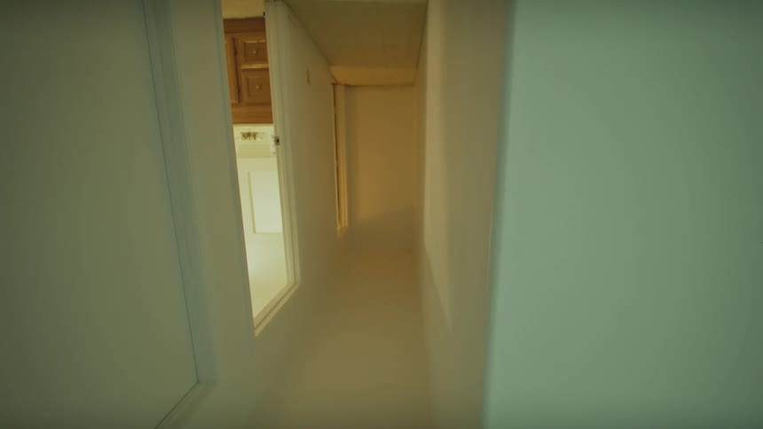 A disorienting, low-angle perspective shot slowly moves through a narrow, white hallway of a house