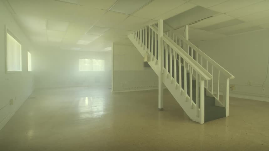 Static shot of an empty, hazy room with a prominent white staircase and dust motes floating in the sunbeams