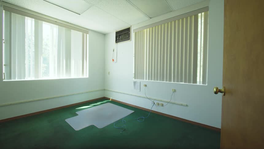 A static shot of a vacant, sunlit office room with green carpet and closed blinds