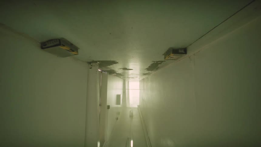 An eerie point-of-view shot moves slowly down a dark, derelict hallway with flickering lights and peeling paint