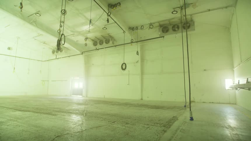 Tilting down from the industrial ceiling to the empty concrete floor of a large, vacant warehouse with flickering green-tinted lights