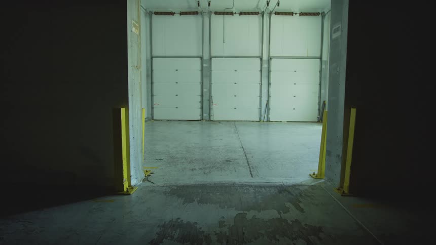 A static shot from a dark hallway looking into an empty, lit warehouse loading dock with three closed garage doors