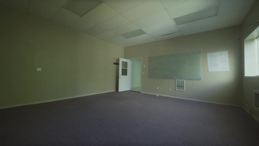A static, wide shot of a vacant office with purple carpet, beige walls, and a whiteboard