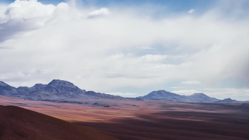 A dramatic timelapse shows clouds and shadows sweeping across a vast, colorful desert mountain landscape