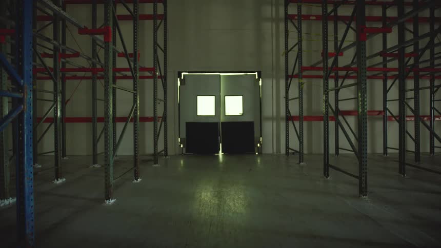 A flashing light from behind a closed double door illuminates the empty shelves of a dark and mysterious warehouse