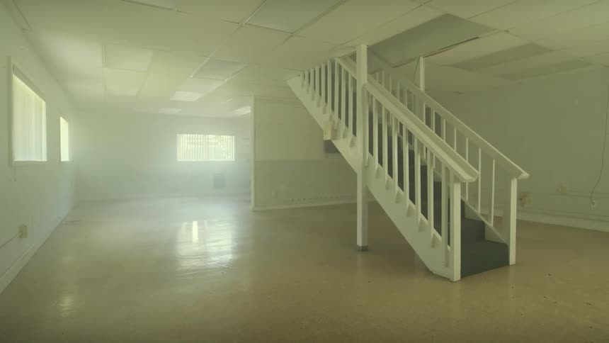 A slow pan reveals an empty, unfurnished room with a central staircase, filled with hazy, bright light