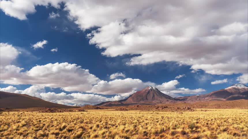 A time-lapse of fluffy white clouds moving across a deep blue sky above a snow-capped volcano in a vast, arid grassy plain