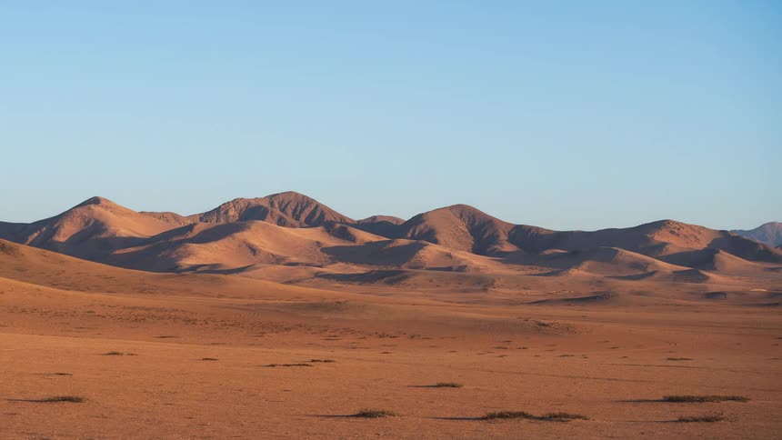 A timelapse of a desert landscape shows the sun setting and casting golden, then purple, hues over the sandy mountains