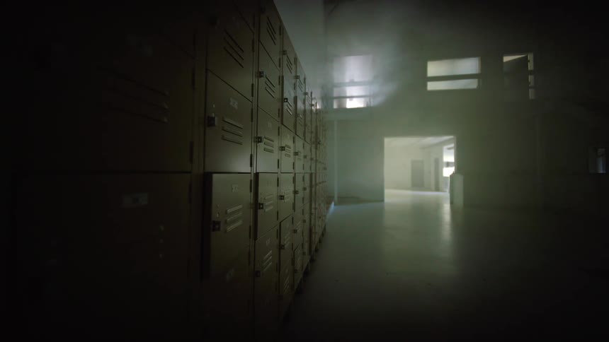 A long row of lockers lines a dark, eerie hallway as smoke slowly drifts through the abandoned building