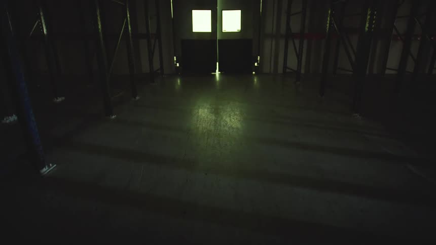 A slow pull-back reveals a vast, dark, and empty industrial warehouse with empty shelving, illuminated only by a distant doorway