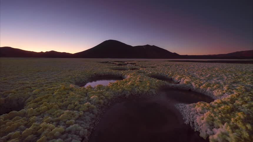 A mesmerizing day-to-night timelapse shows the sun setting over surreal yellow mineral deposits and water pools in a vast salt flat, revealing a brilliant starry sky