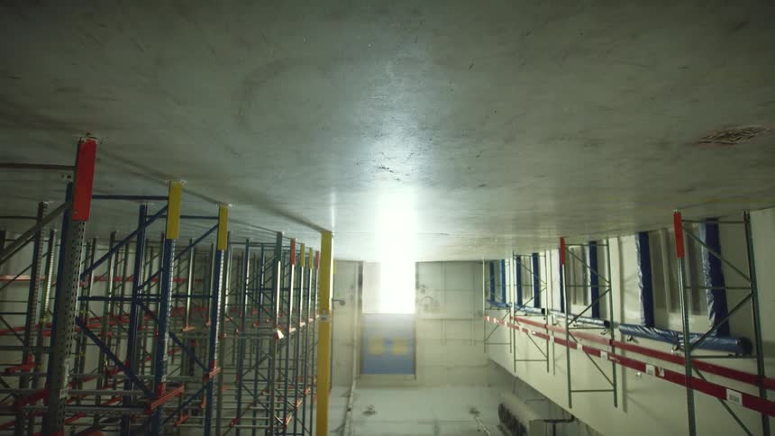 A slow upward crane shot reveals the empty interior of a massive industrial warehouse with tall, colorful shelving racks