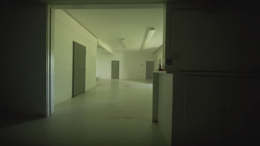 An eerie dolly shot moves through a deserted, sterile institutional hallway with multiple closed doors and dim fluorescent lighting