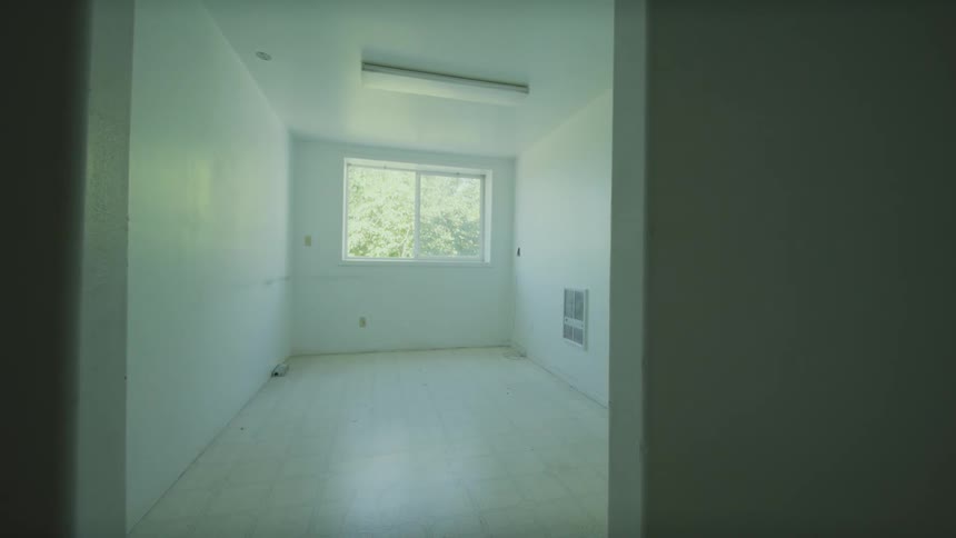 A slow dolly out shot from a small, empty, white-walled room with a window revealing the interior of a vacant house