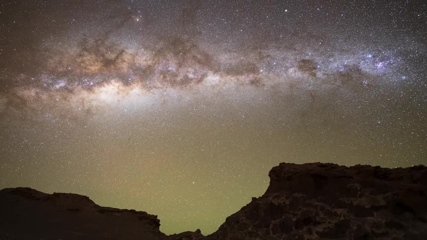 A breathtaking time-lapse captures the Milky Way galaxy and countless stars rotating across the night sky, transitioning to dawn above desert rock formations