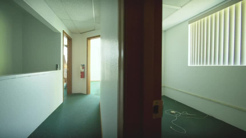 A slow tracking shot moves backwards through a vacant office hallway, revealing an empty, unfurnished room with a window