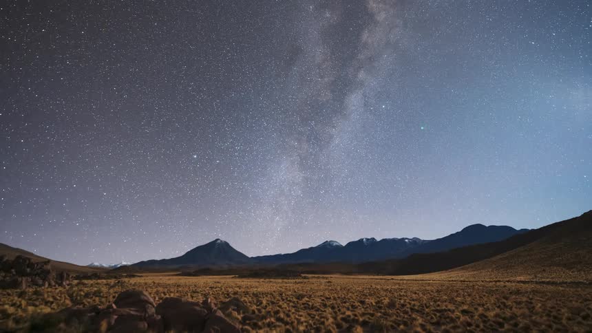 An epic night-to-day time-lapse captures the colorful Milky Way galaxy transitioning to a brilliant sunrise over a vast, arid mountain valley