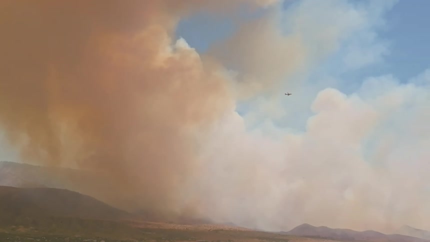 An aircraft bravely navigates through massive plumes of thick, dark smoke rising from a raging wildfire in the mountains