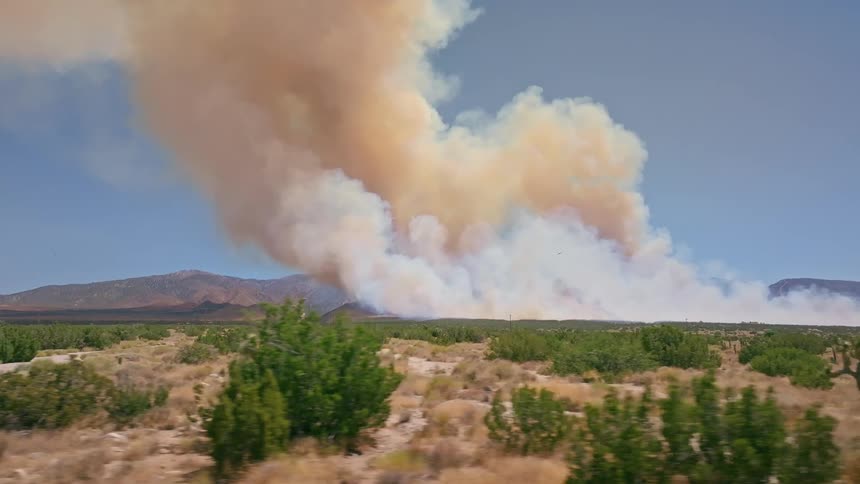 Aerial dolly shot across the Mojave Desert reveals a massive wildfire sending a huge plume of orange and white smoke into the blue sky