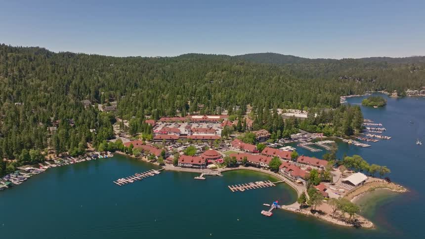 An expansive aerial shot slowly moves toward a scenic lakeside resort town with a marina, surrounded by lush green forests on a bright day