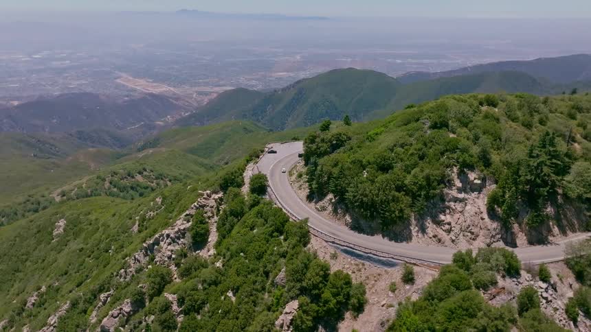An aerial pullback reveals cars navigating a hairpin turn on a scenic mountain road, with a sprawling cityscape visible in the distance