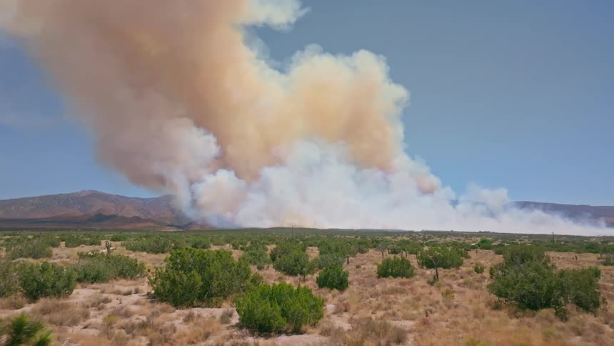 A slow dolly shot moves through the Mojave Desert landscape, revealing a massive wildfire and a huge plume of smoke on the horizon