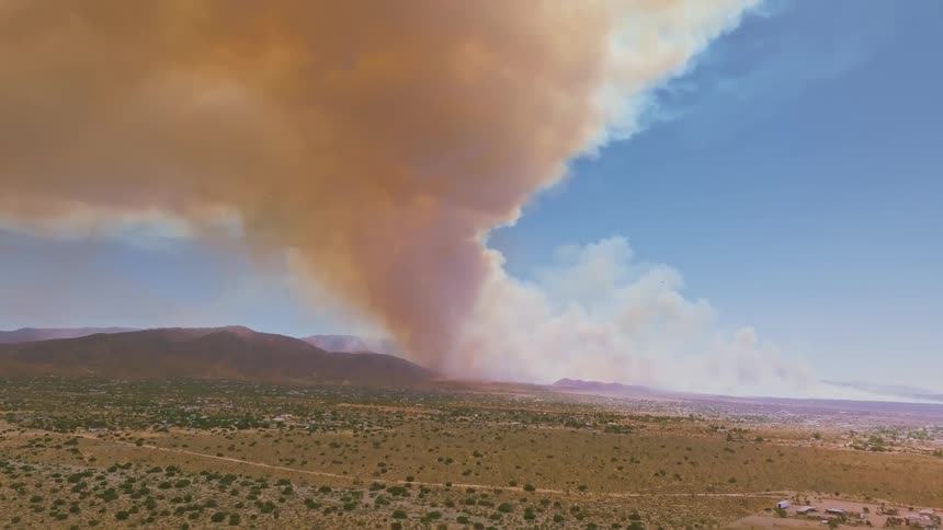 An enormous wildfire rages in the mountains, sending a massive plume of thick brown smoke billowing into the blue sky over a desert landscape