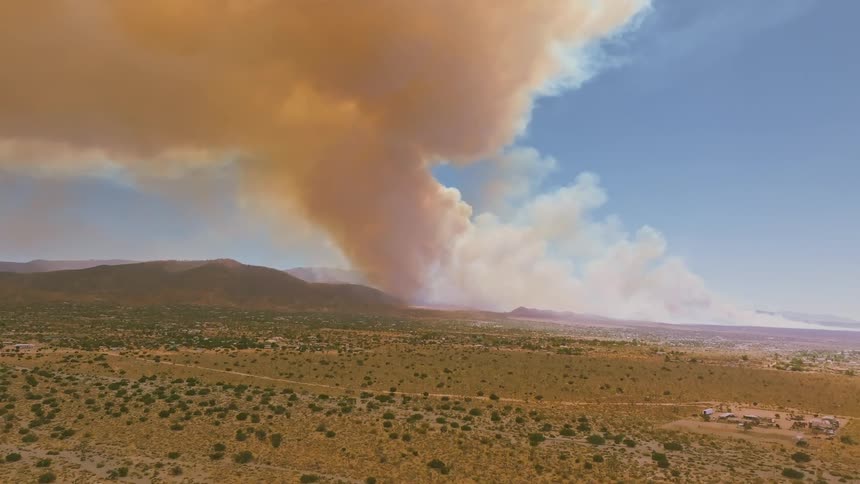An aerial view shows a massive wildfire sending a huge plume of thick smoke into the sky over a dry desert landscape