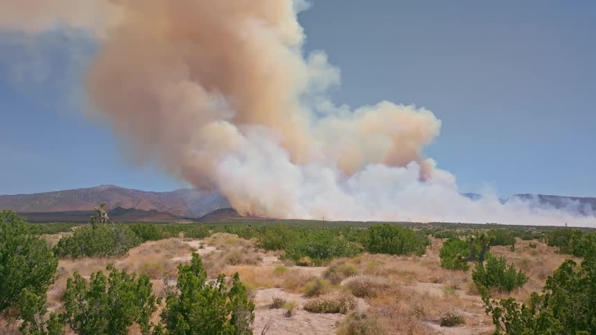 A massive plume of orange and white smoke billows into the blue sky from a wildfire burning through a dry, brushy landscape with mountains in the background