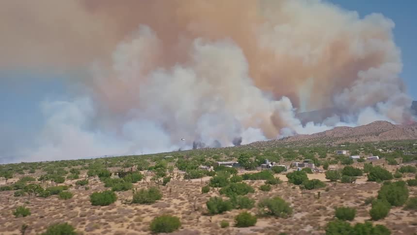 An aerial shot pulls back to reveal a massive wildfire burning in the desert, with a huge column of smoke filling the sky and a helicopter flying to the scene