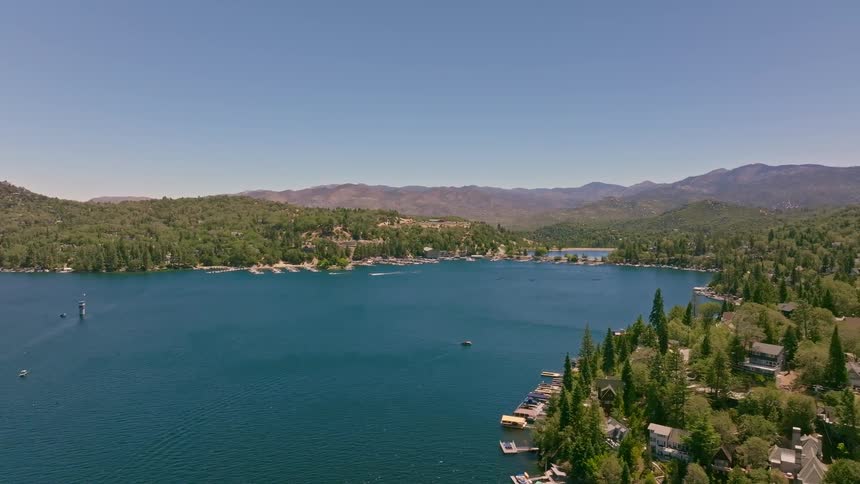 A beautiful aerial shot slowly flying over a vast mountain lake surrounded by lush green forests and homes on a sunny day