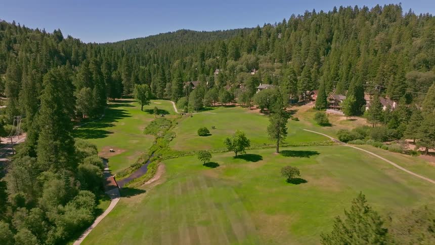 A scenic aerial journey glides over a beautiful golf course tucked away in a sunlit valley surrounded by a dense pine forest