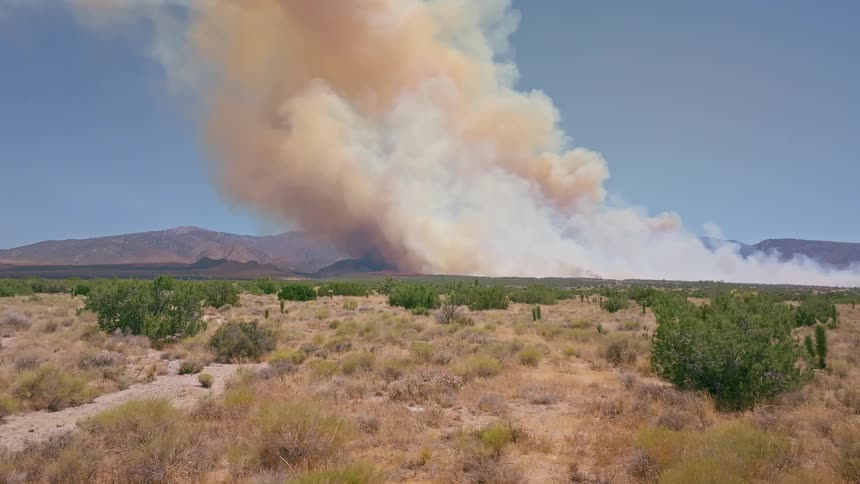 A massive wildfire sends a huge plume of smoke into the sky over a dry desert landscape