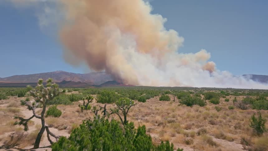 An aerial view pushes in over the Mojave Desert, revealing a massive wildfire sending a huge plume of smoke into the sky from the nearby mountains