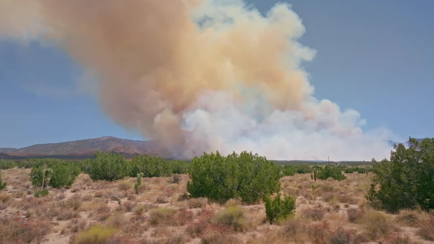 A slow sideways aerial reveals a massive wildfire sending a huge plume of thick smoke into the sky over a dry desert landscape
