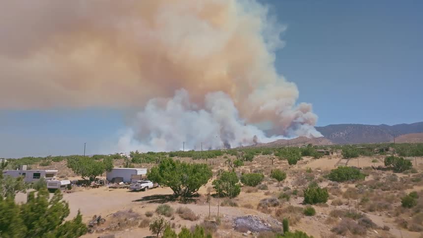 A slow aerial view pushes over a dry desert landscape towards a massive wildfire sending a huge plume of smoke into the sky