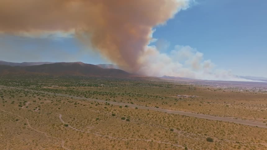 A dramatic aerial view shows a massive wildfire sending a huge column of thick smoke into the sky over a vast desert landscape