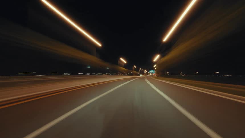 Point-of-view hyperlapse of driving down an illuminated city highway at night, creating a mesmerizing light trail and blur effect