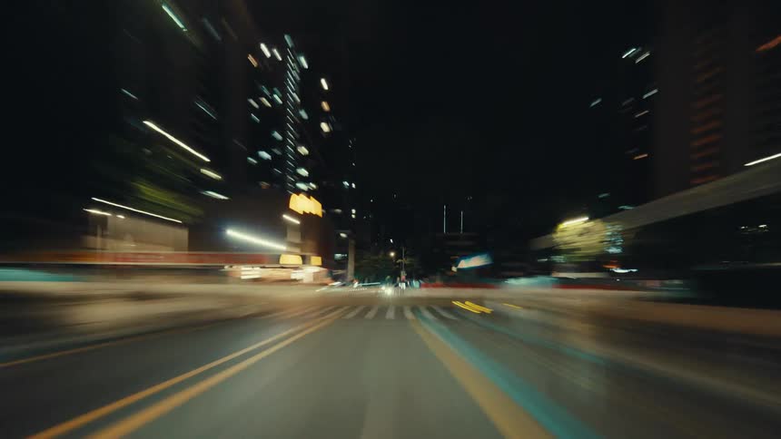 A dynamic hyper-lapse captures a first-person view of driving through a bustling city at night, with vibrant light trails streaking past