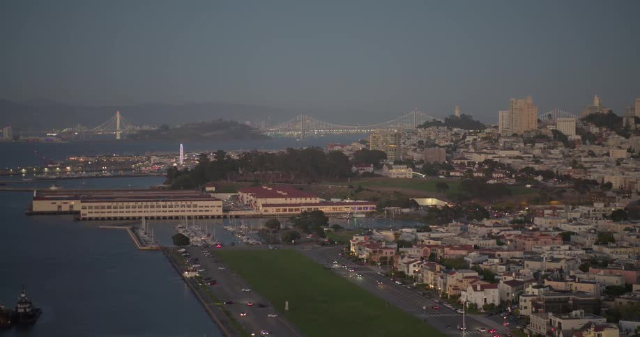 A scenic wide shot of the San Francisco harbor and Marina Green at dusk with the Bay Bridge illuminated in the distance
