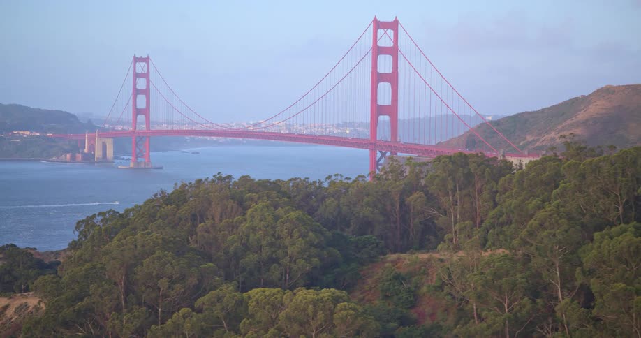 A scenic tracking shot reveals the iconic Golden Gate Bridge spanning the San Francisco Bay at dusk, with lush green forested hills in the foreground