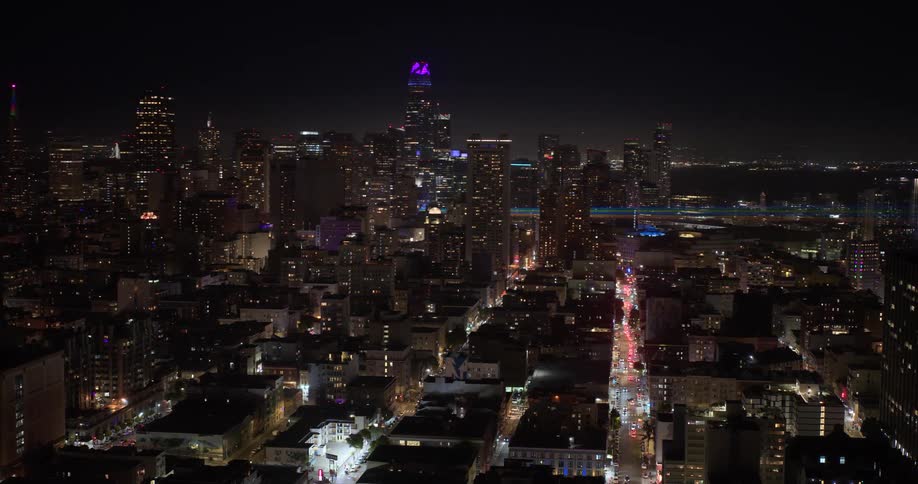 An aerial view reveals the glowing San Francisco skyline at night, with the Salesforce Tower and Bay Bridge illuminated in the distance