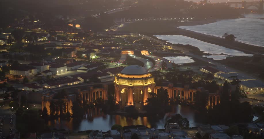 A stunning high-angle, slow zoom-out shot of the illuminated Palace of Fine Arts in San Francisco at twilight, revealing the cityscape and bay