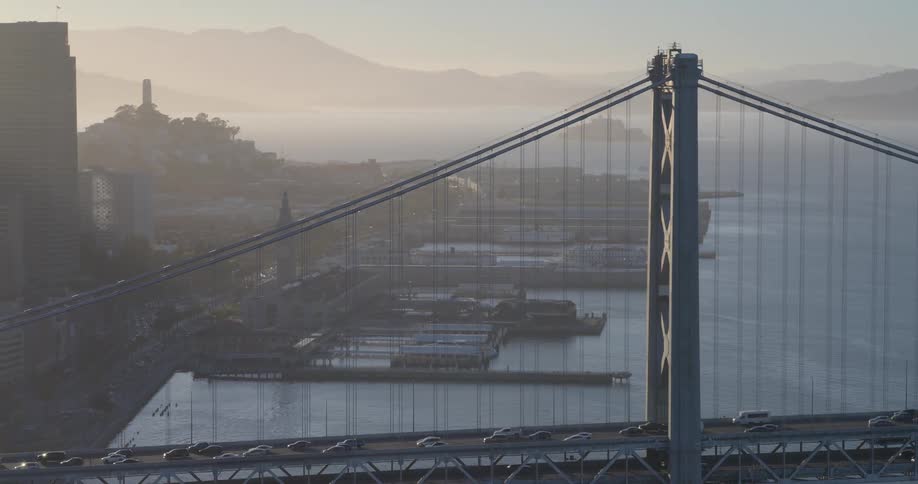 Slow aerial pull-back reveals the iconic San Francisco skyline behind the Bay Bridge as traffic flows during a hazy golden hour