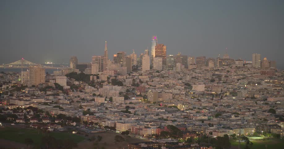 A wide, static shot of the iconic San Francisco skyline with the Bay Bridge visible in the distance as the city lights illuminate the twilight sky